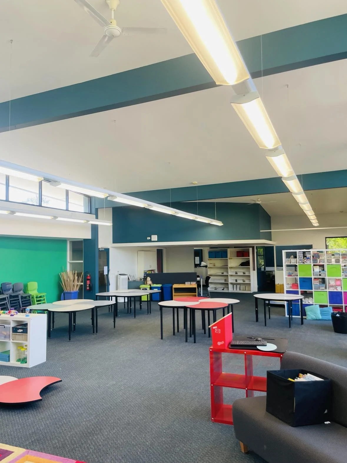 Interior of a classroom or activity room with tables, colorful storage shelves, chairs, and educational materials, illuminated by ceiling lights and large windows.
