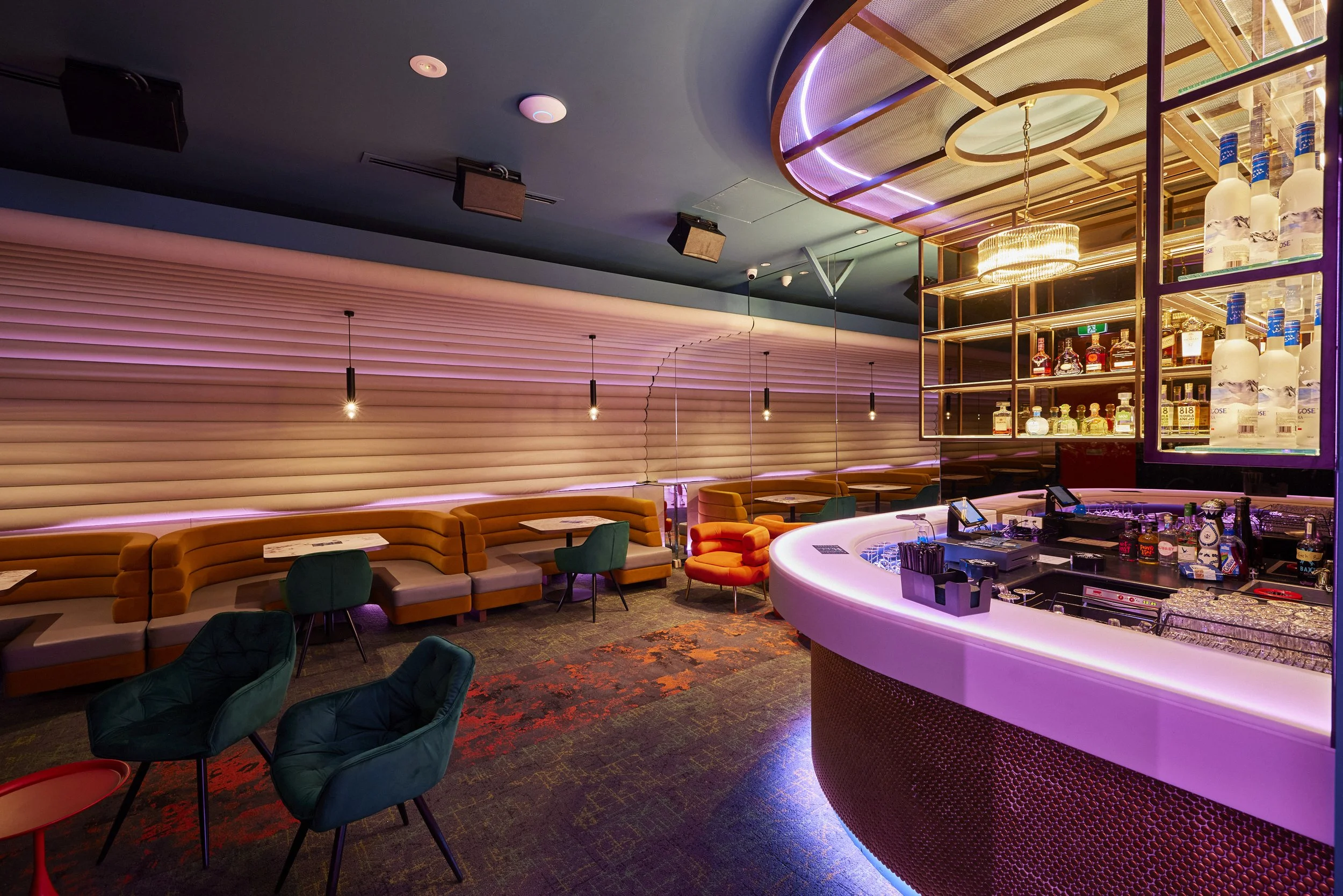 A modern bar with colorful furniture, soft lighting, and a well-stocked liquor shelf, featuring neon accents and contemporary décor.