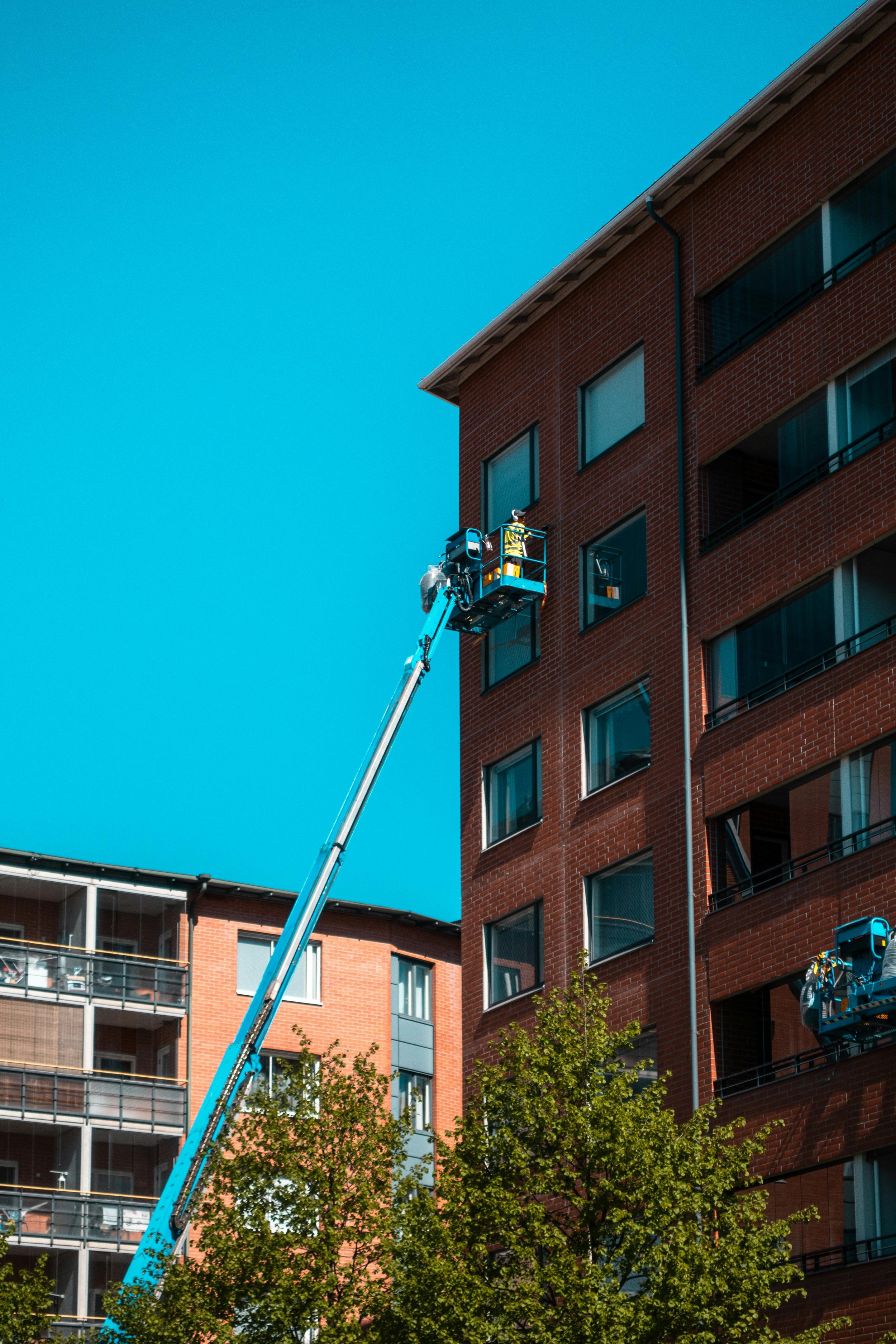 A worker in a yellow safety vest operating a cherry picker near a multi-story red brick building with green trees in the foreground and a clear blue sky in the background.