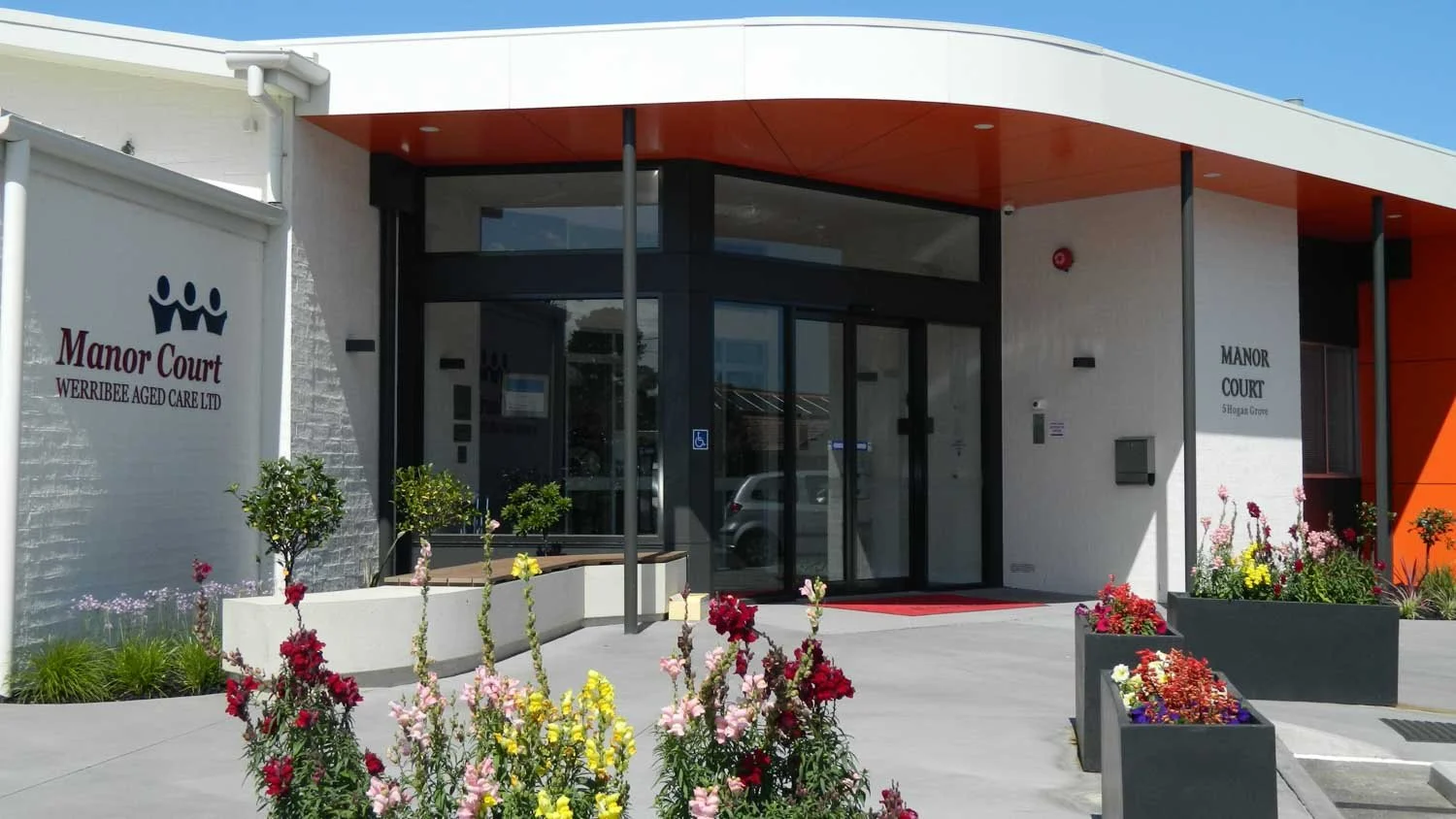 The entrance to Manor Court, a care facility with glass doors, potted plants, and colorful flowers outside, under a white and orange awning.