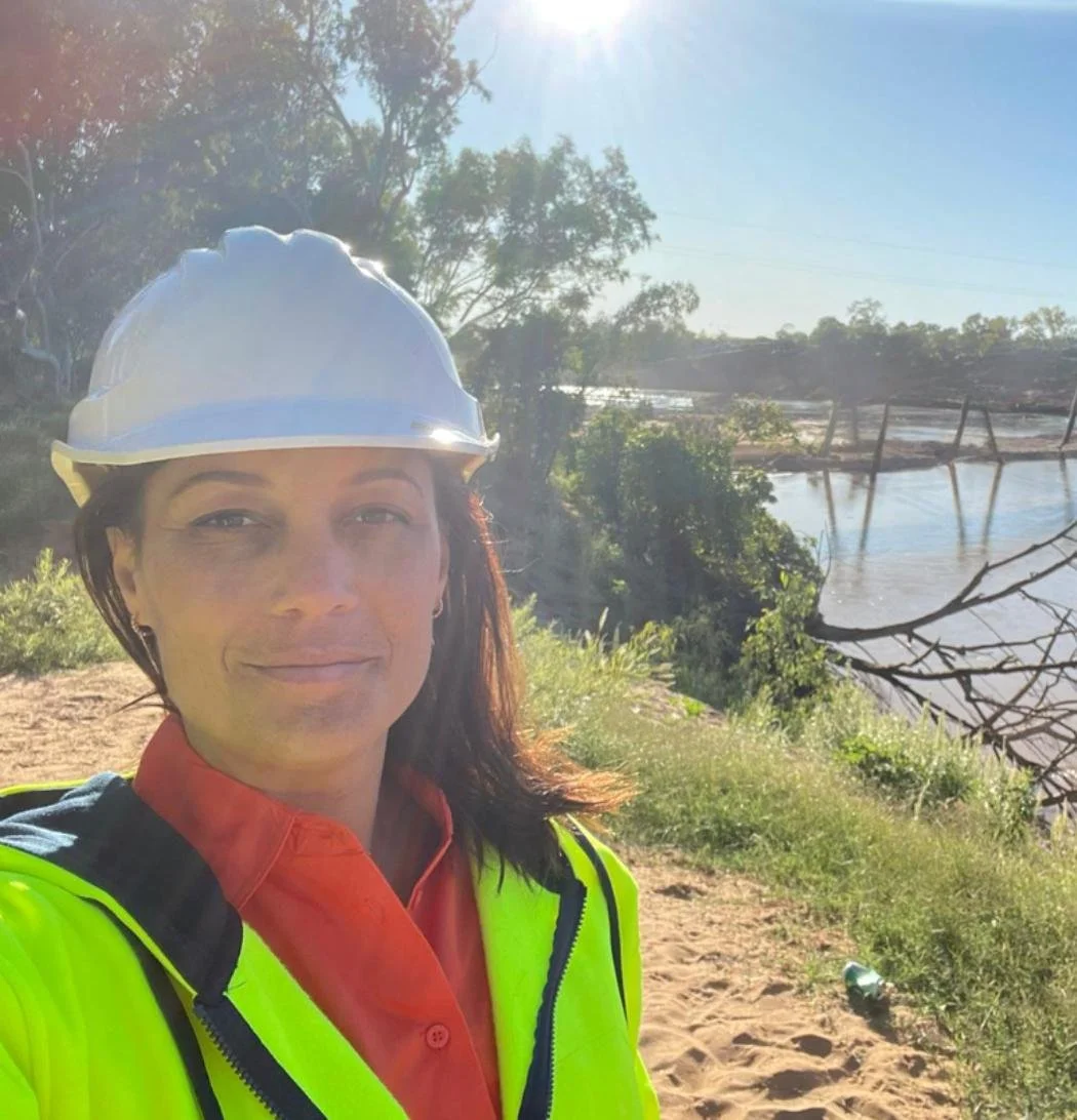 A woman wearing a white hard hat and a neon safety vest taking a selfie outdoors near a river or body of water, with trees and a clear sky in the background.