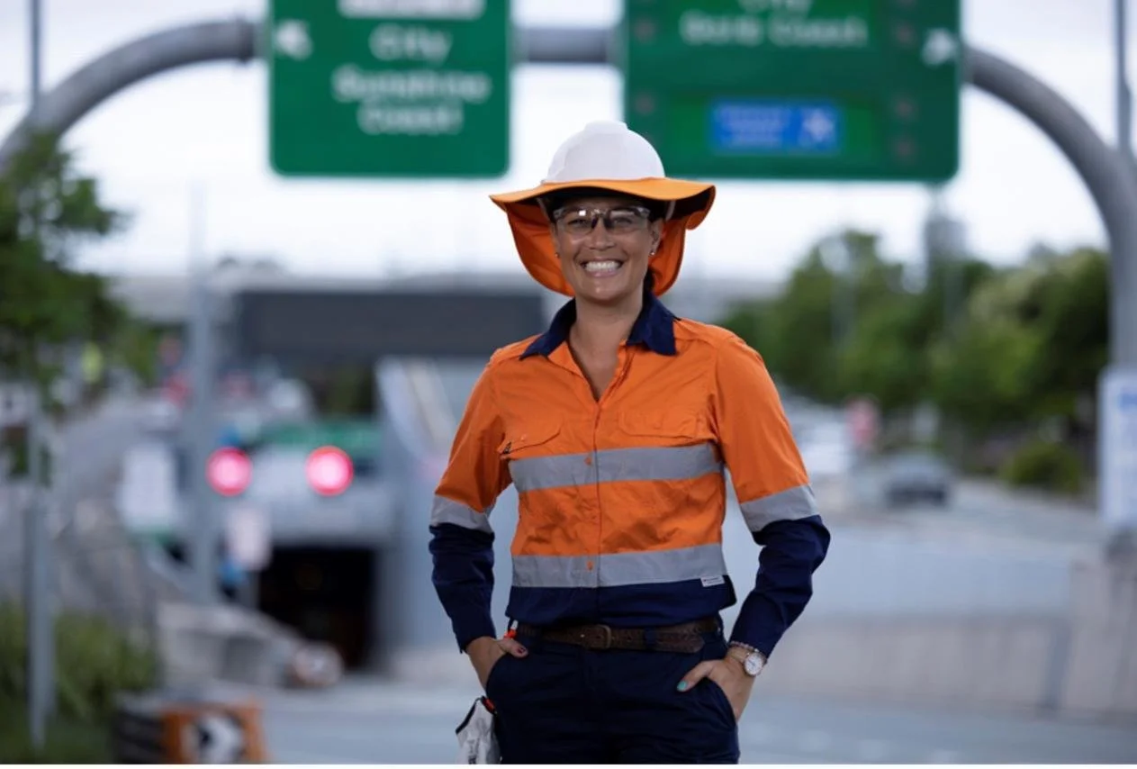 A smiling female construction worker wearing an orange safety jacket, a white and orange safety helmet, and safety glasses, standing with her hands on her hips in front of a highway with cars and green road signs.