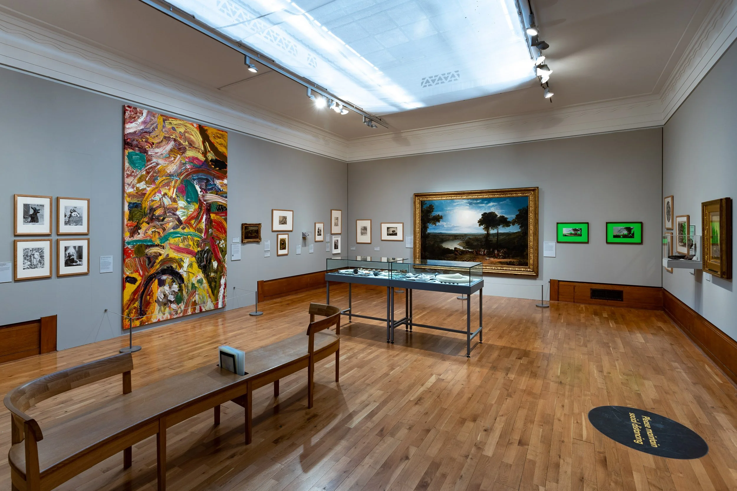An art gallery room featuring various artworks, including paintings on the walls and a large, colorful abstract painting. There is a glass display case with jewelry or small sculptures on a wooden floor, and a doorway leading to another exhibition space.