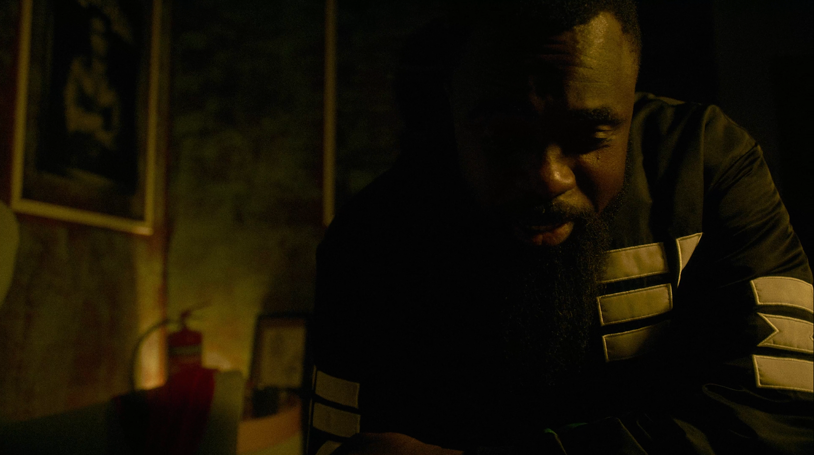 A man with a beard and mustache in dark lighting, leaning forward with eyes closed or looking down, with a fire extinguisher and framed picture in the background.