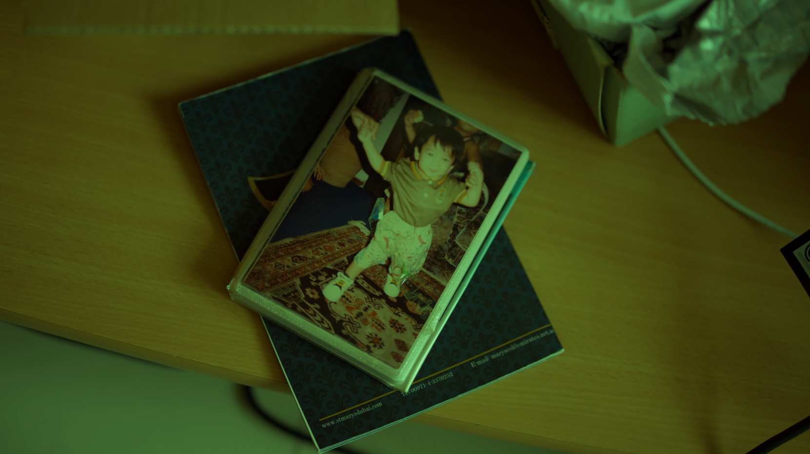 Child standing on a patterned carpet, raising arms in a pose, inside a clear rectangular case, placed on a surface with a book underneath and a crumpled paper bowl nearby.