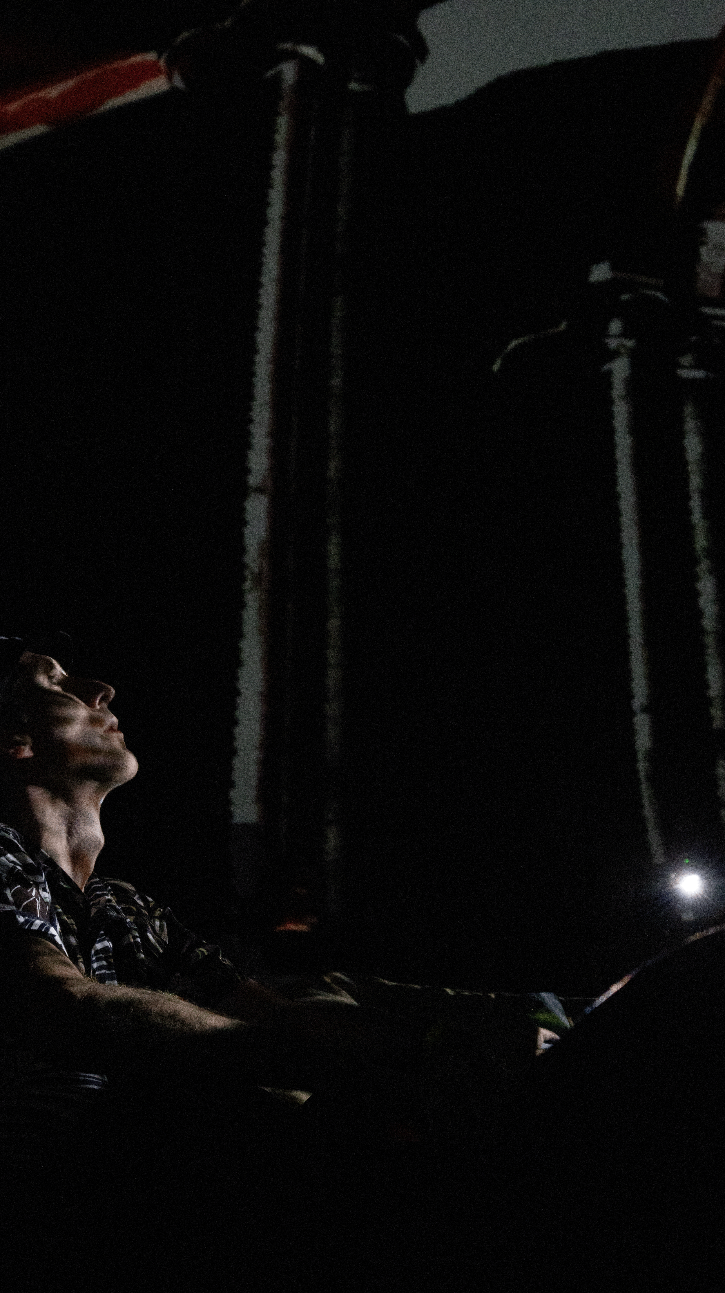 A person with short hair wearing a patterned shirt is sitting in a dark room, illuminated by a small light, with tall dark structures in the background.