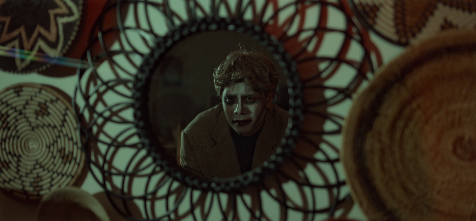 A young man with disheveled hair and dark makeup around his eyes looks into a small circular mirror, surrounded by various woven baskets and decorative wall hangings.