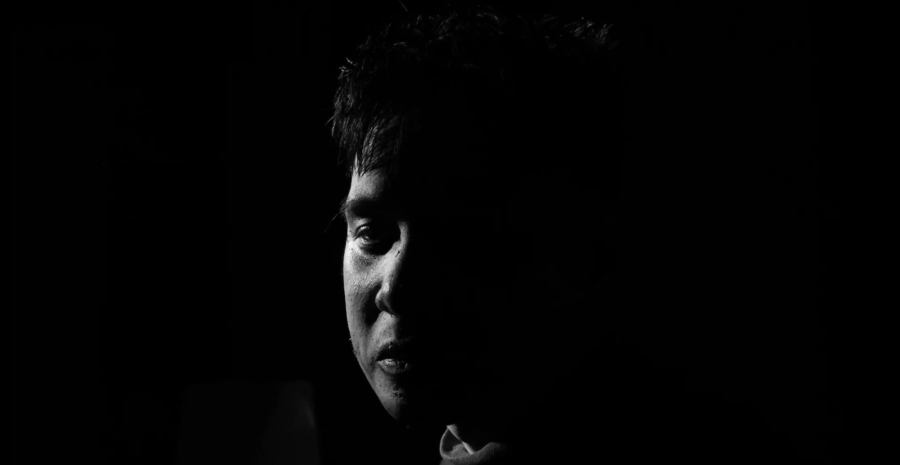 A black and white portrait of a person with short hair, partially illuminated on one side of their face, with the rest in shadow.