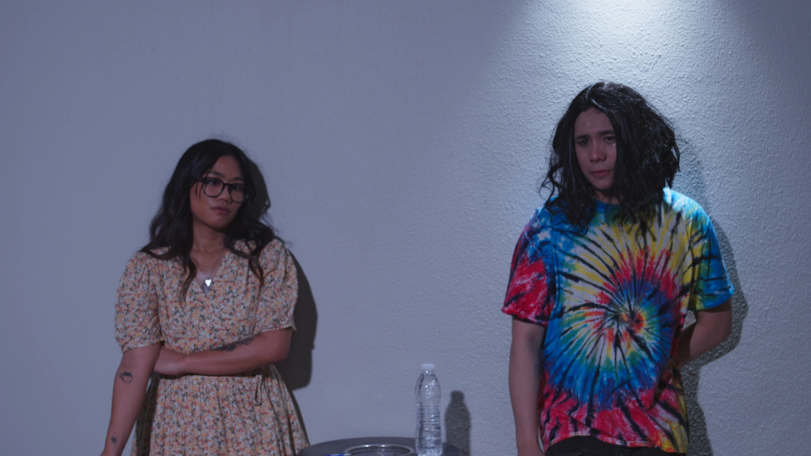 Two young women stand against a plain gray wall, one with glasses and tattoos, wearing a floral dress, and the other with curly hair wearing a colorful tie-dye shirt, both looking somber.