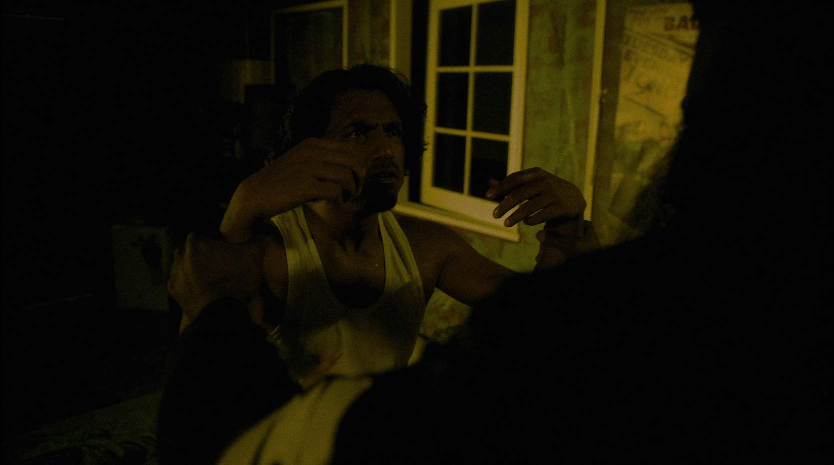 A man with dark hair and beard wearing a yellow tank top, sitting in a dimly lit room, gesturing with his hands, with a window and a map on the wall behind him.