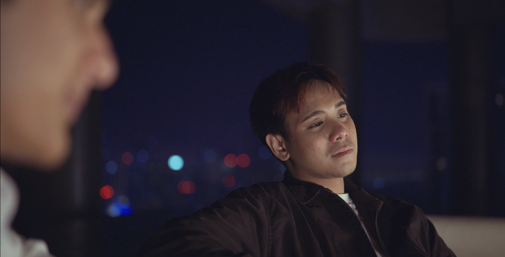 A young man looking thoughtfully with city lights blurred in the background during nighttime.