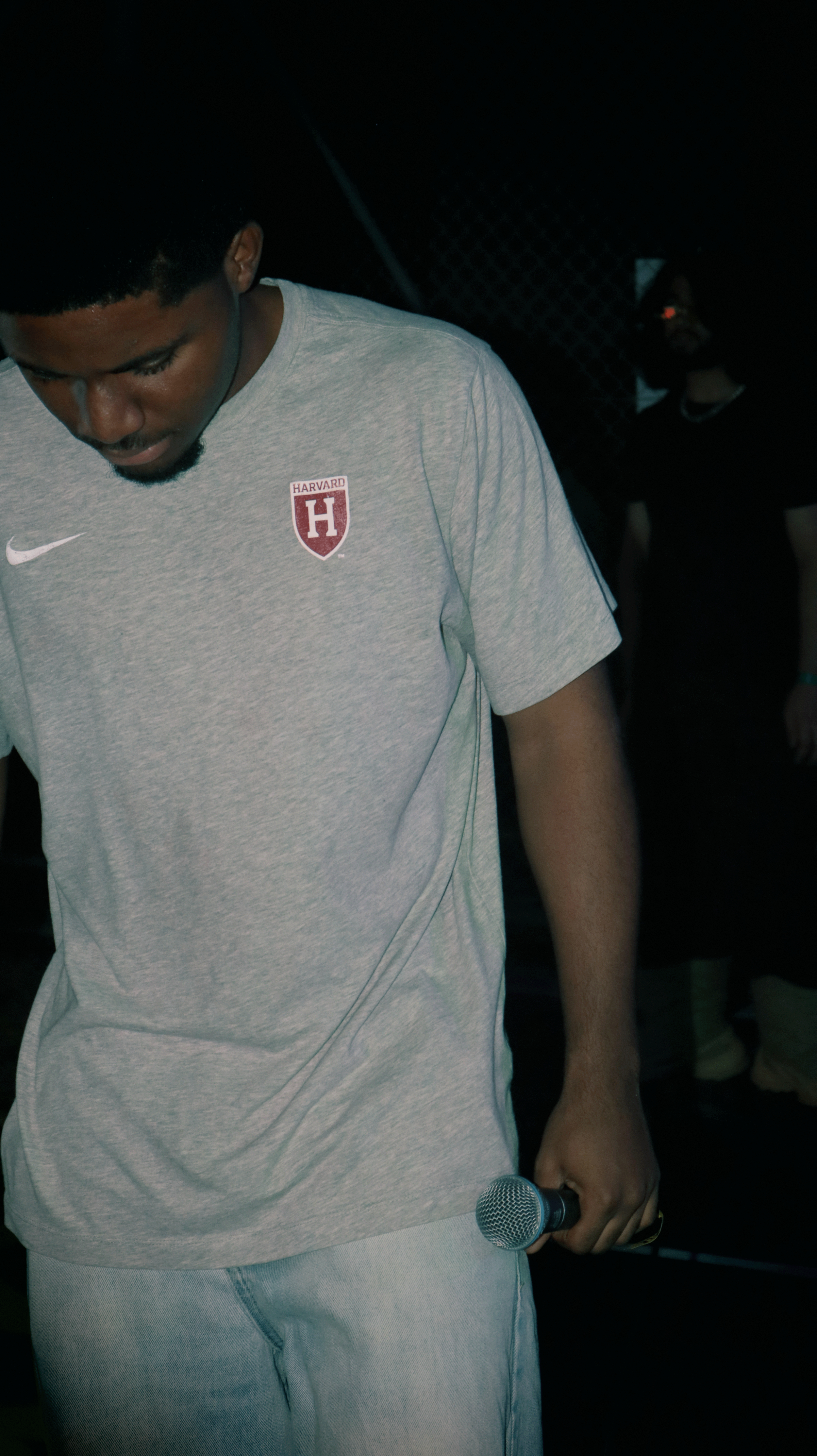 Young man holding a microphone, wearing a light gray Harvard shirt, looking down, in a dark environment