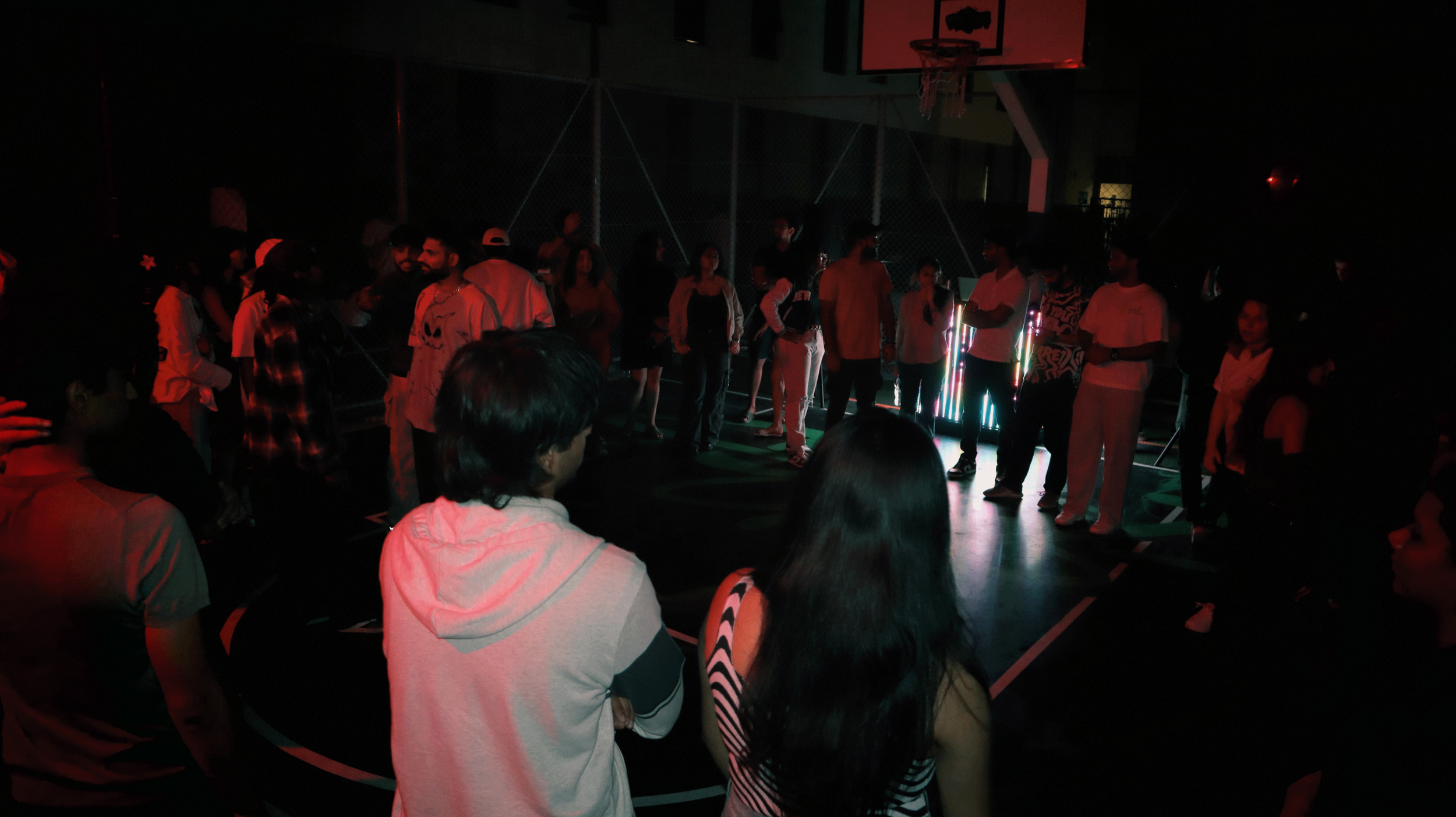 Group of people standing in a dark, illuminated gymnasium or sports hall, possibly during a party or event, with minimal lighting and some decorative lights.
