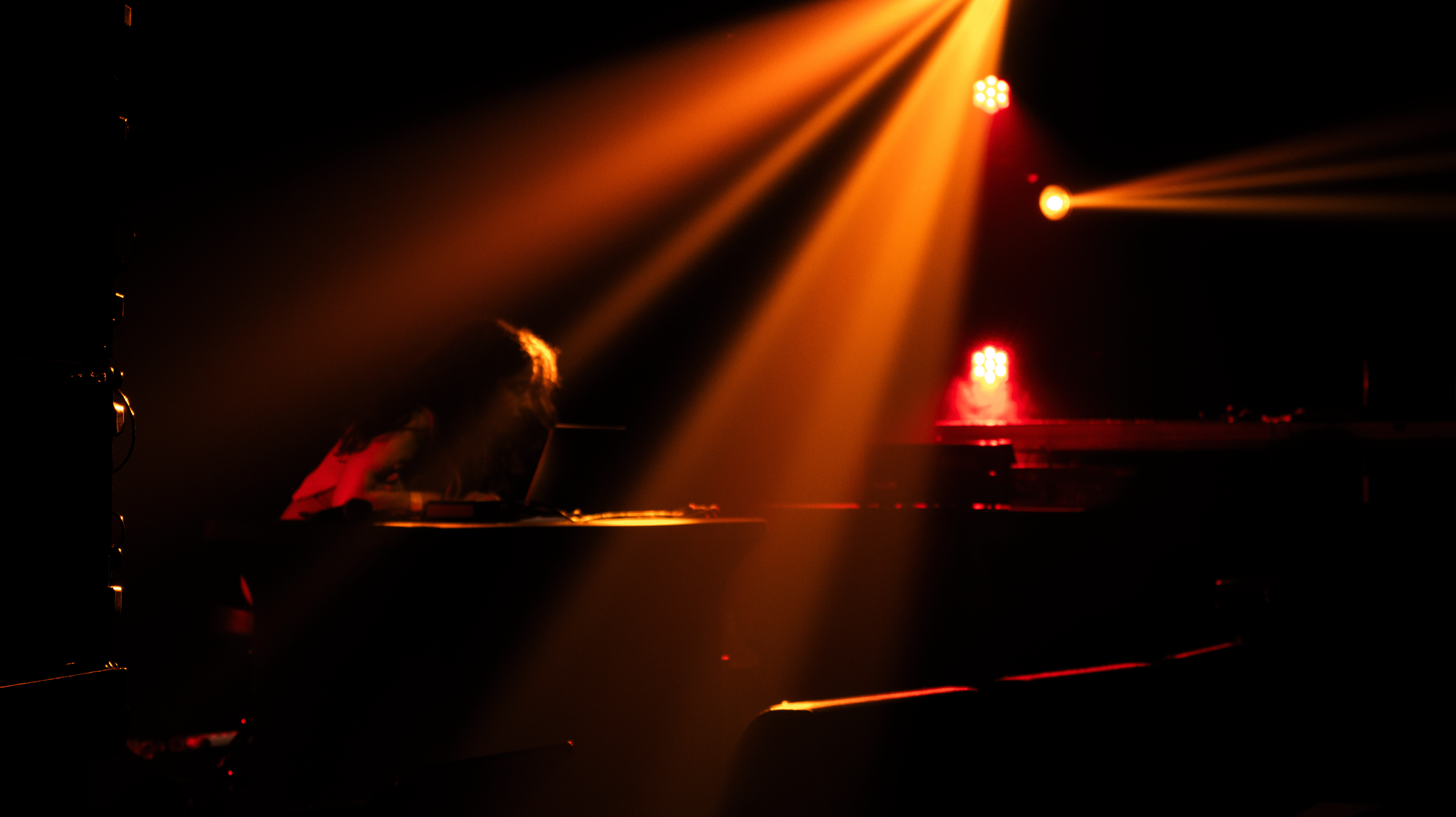 A person with long hair sitting at a piano under stage lighting with orange and red spotlights.