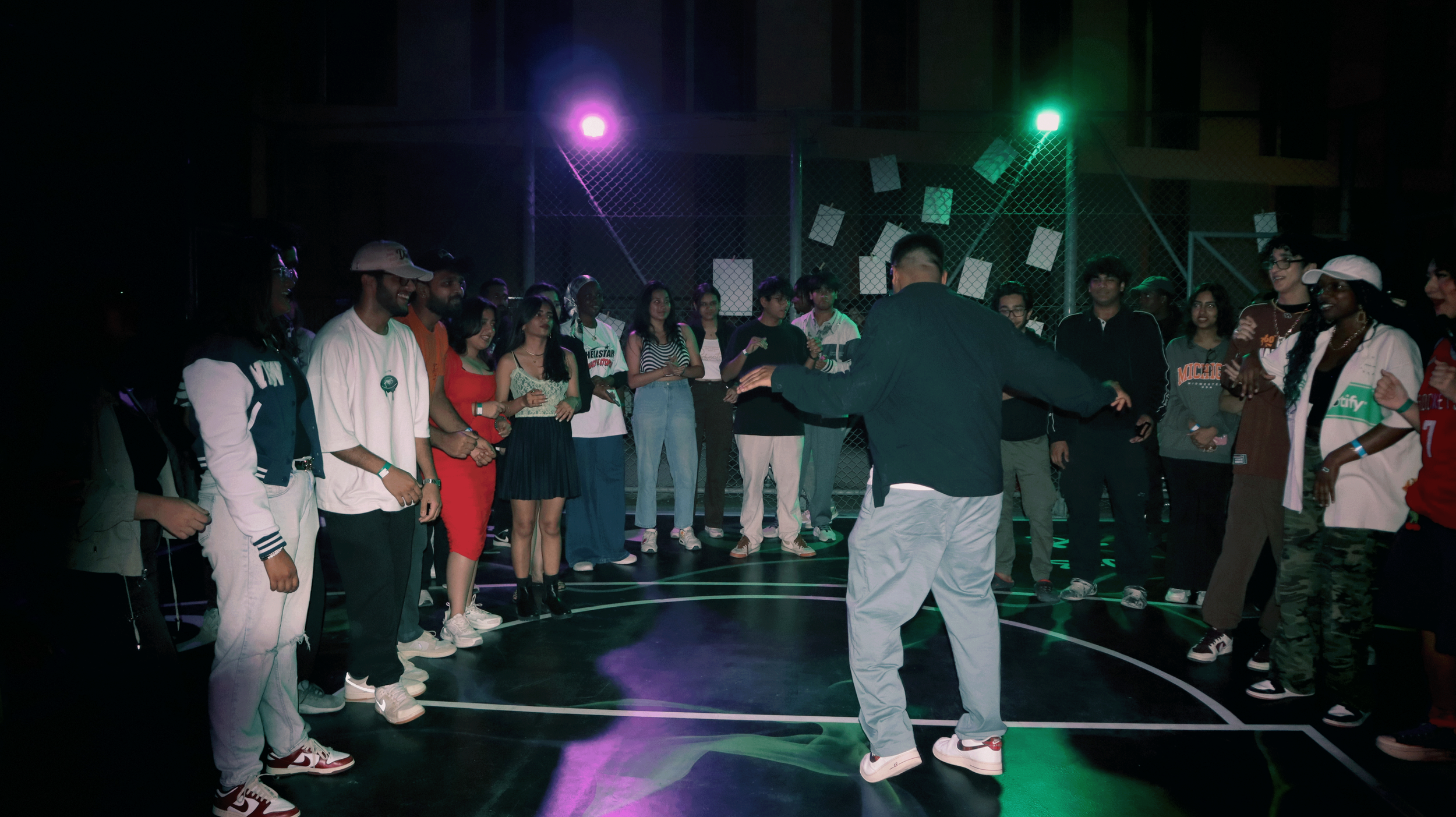 Group of people gathered on a basketball court at night, listening to a man in a black jacket and gray pants speak or perform, with colorful lights illuminating the scene.