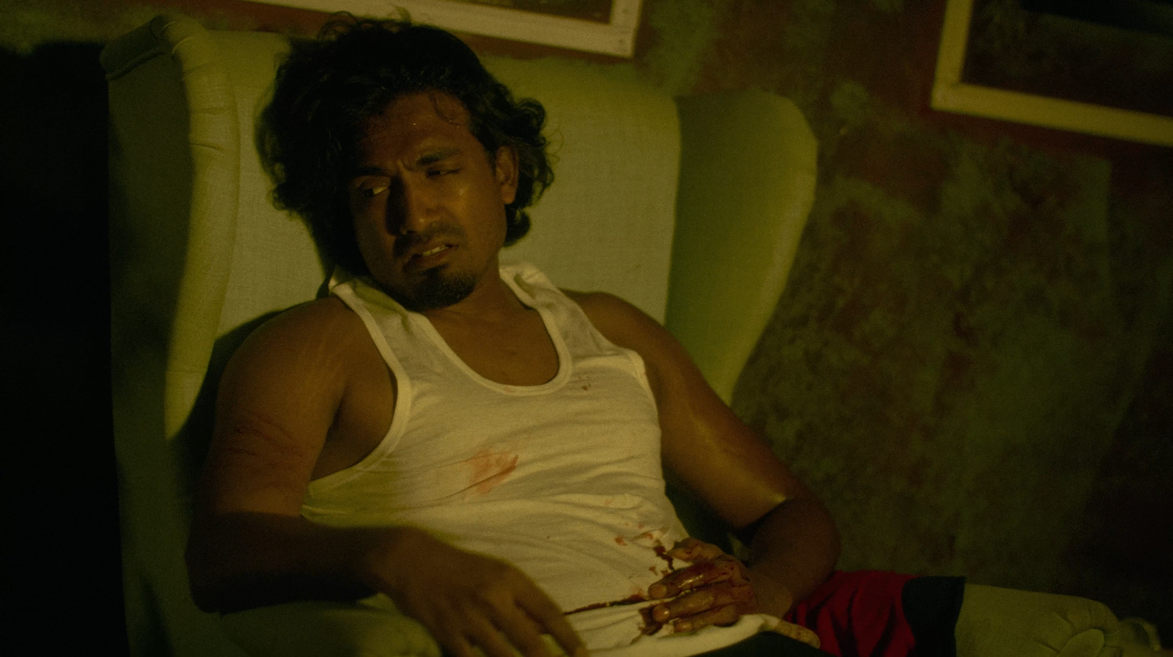 A man with curly black hair and a beard lying on a bed, wearing a white tank top stained with blood, holding a knife with blood on it, looking distressed in a dimly lit room.
