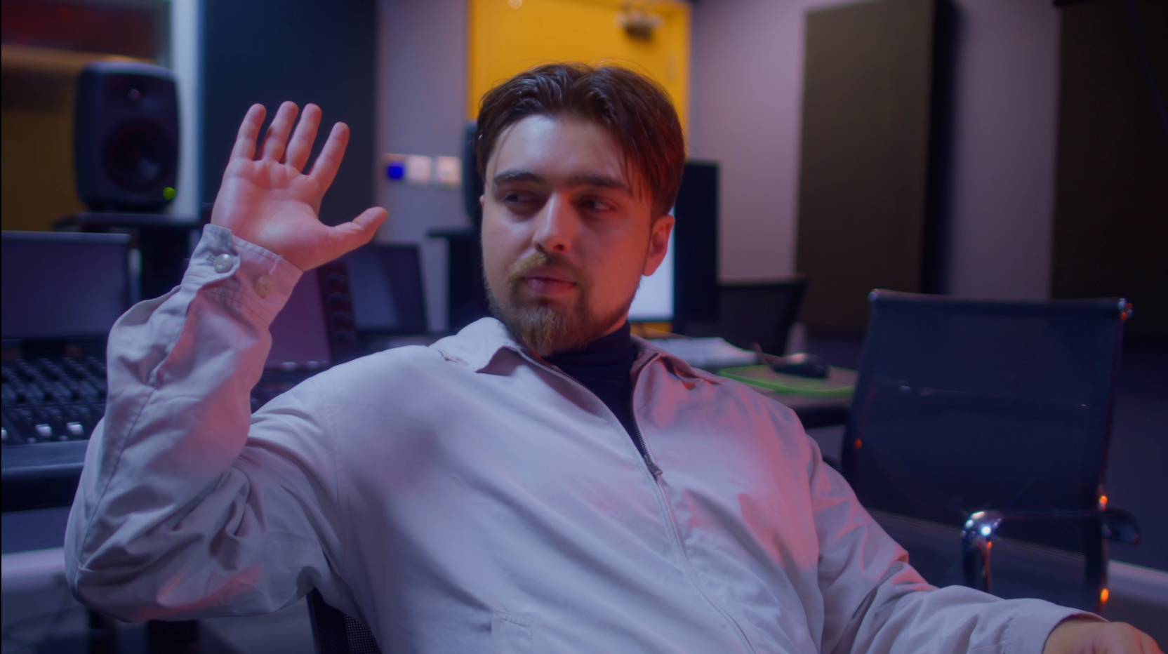 A man with a beard and brown hair, wearing a white jacket and black shirt, is seated in a recording studio with sound equipment. He is raising his right hand with a relaxed expression, looking to his left.
