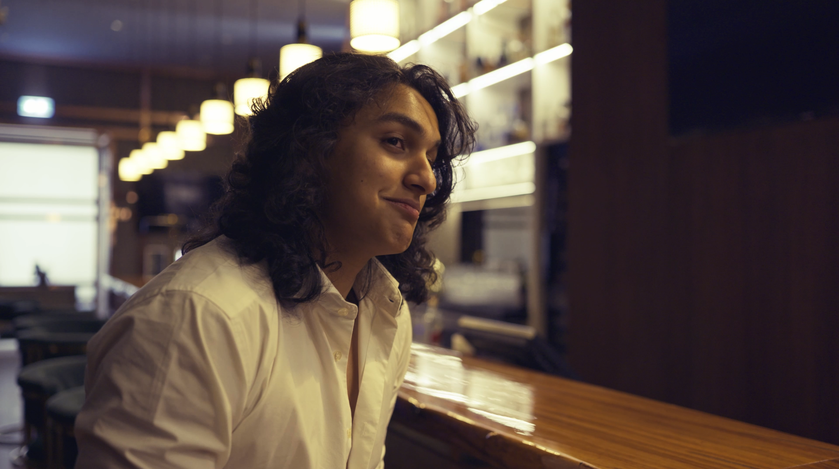 A person with dark, curly hair wearing a beige button-up shirt, sitting at a bar counter inside a dimly lit space with warm overhead lights and shelves with bottles in the background.