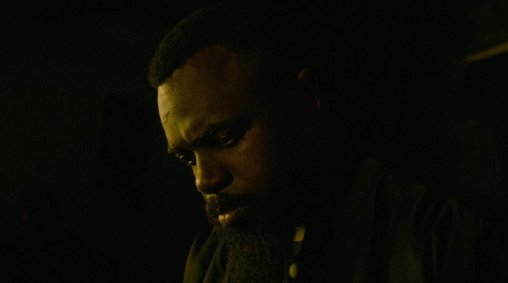 A man with a beard and short hair, looking down in a dark environment with low lighting.
