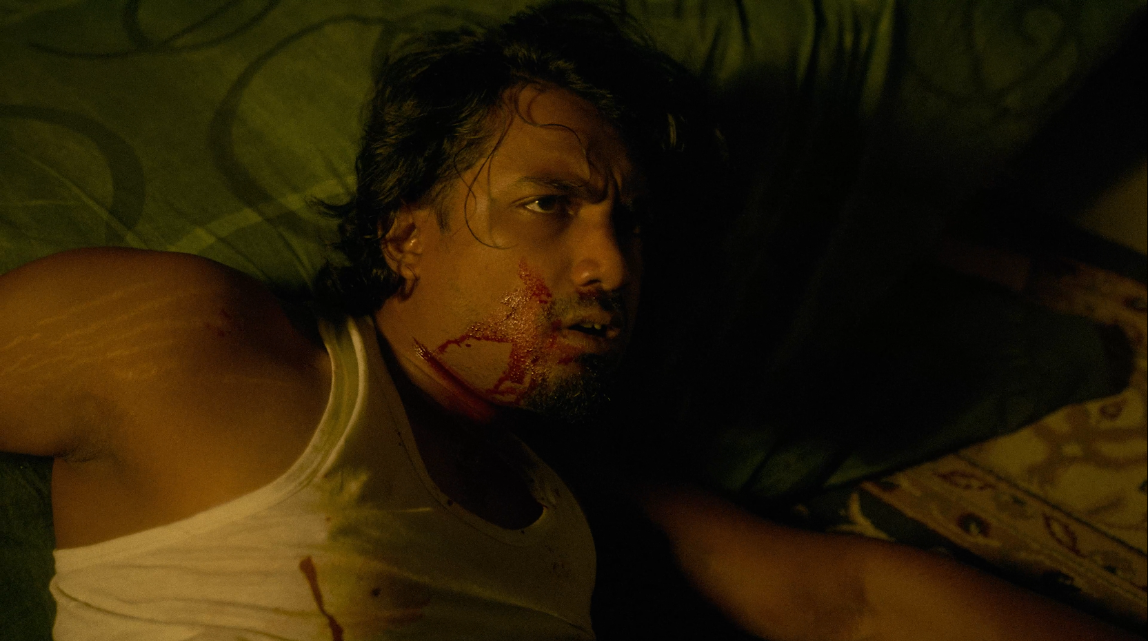 A man with long dark hair lies on a dark green pillow, with blood on his face and a bloodstain on his white tank top. The lighting is dim, giving the scene a tense or dramatic atmosphere.