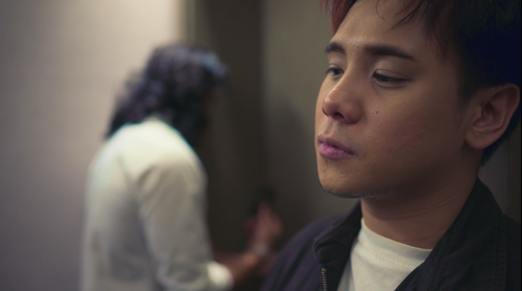 A young man with short dark hair and a somber expression is leaning against a wall, while a blurred woman with dark hair and a white shirt is in the background.