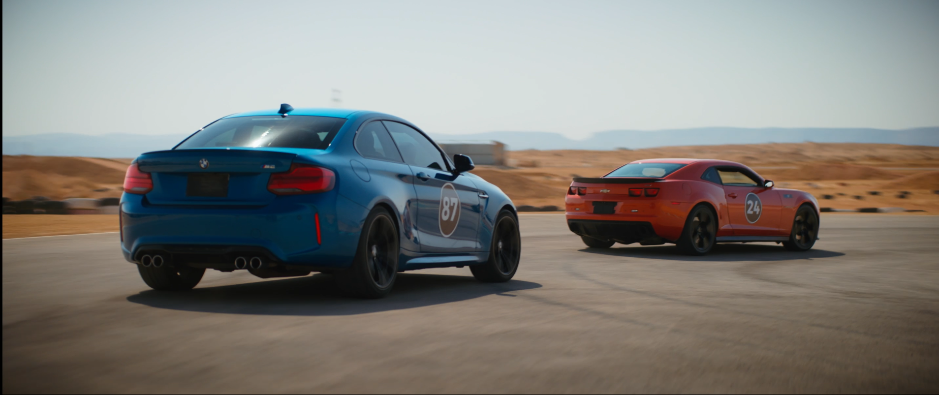 Two cars, a blue BMW and a red Chevrolet, racing on a track in a desert landscape with hills in the background.