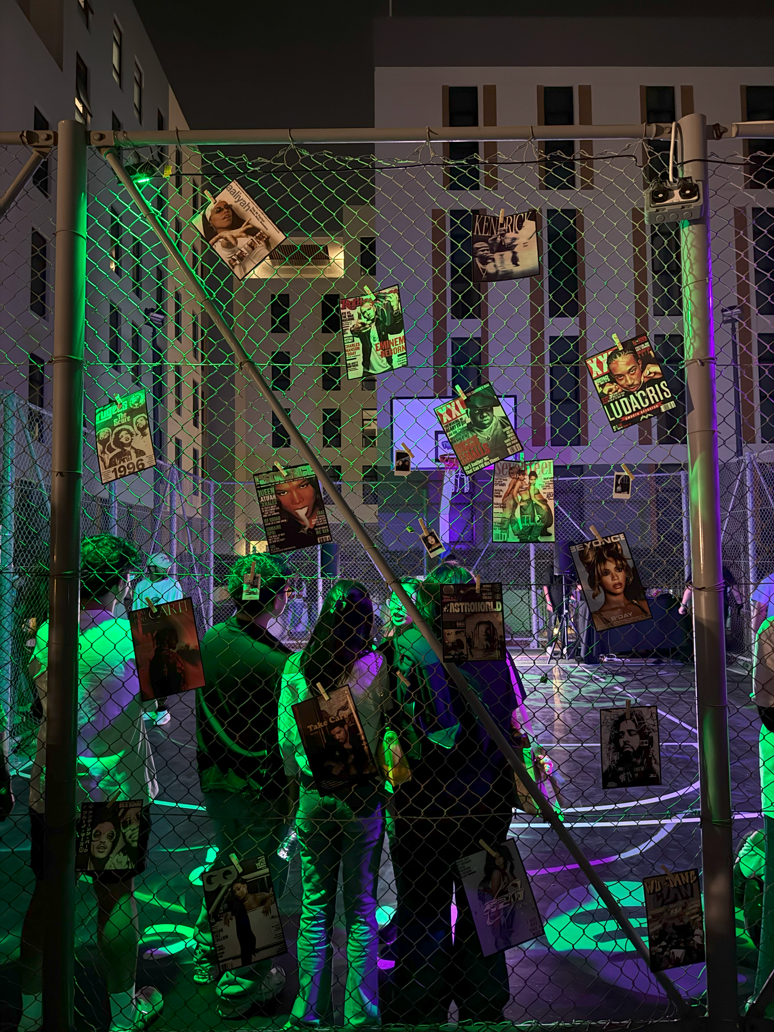 Group of people standing behind a chain-link fence decorated with magazine posters in a nighttime setting with colorful neon lights.