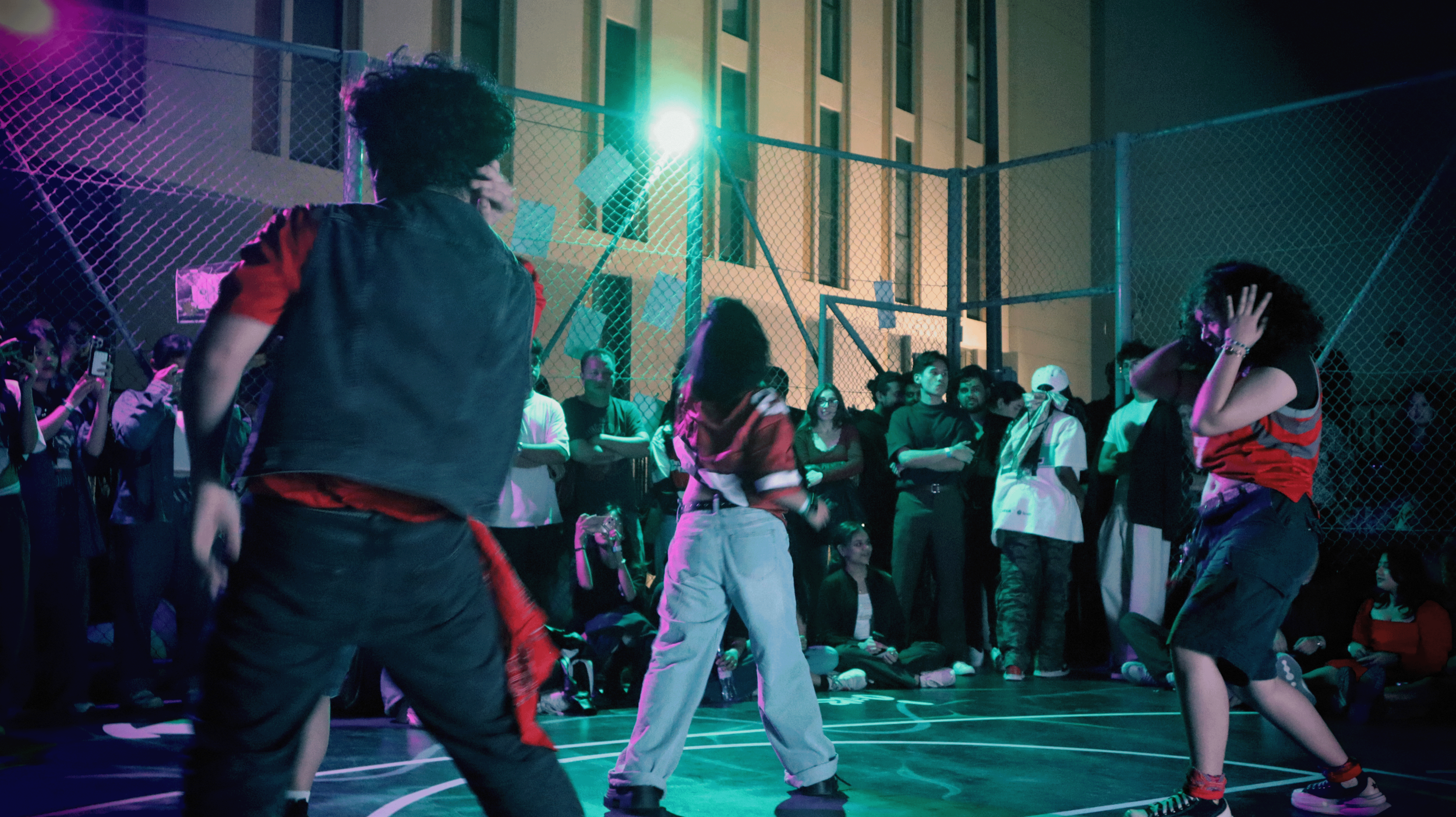A group of young people dancing on a rooftop basketball court at night, with a crowd watching them and a building in the background, lit by colorful neon lights.