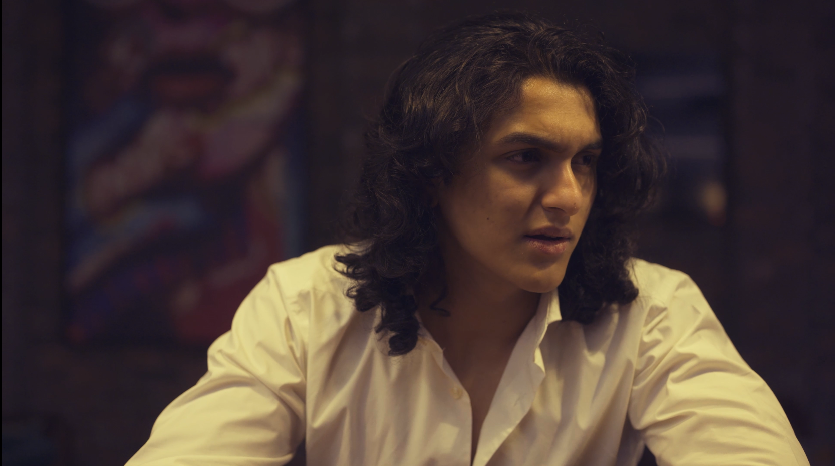 Young man with long curly dark hair and a serious expression, wearing a white button-up shirt, sitting indoors with a dark background and abstract art on the wall.