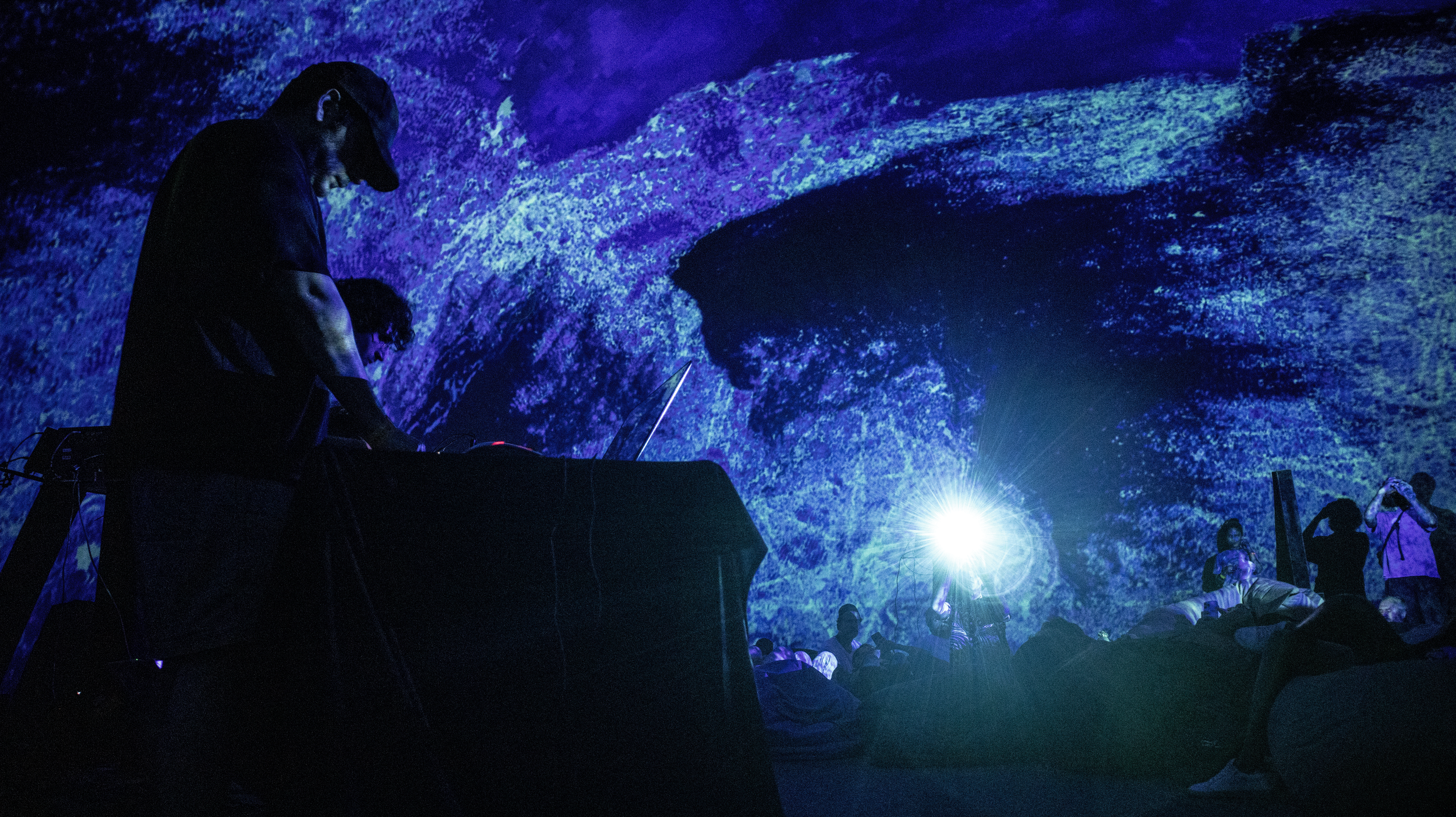 A live electronic music performance in a dark room with a blue and purple digital projection on the wall. A DJ stands at a table with electronic equipment, and a crowd of people is sitting or lying on the floor, some taking photos or videos.