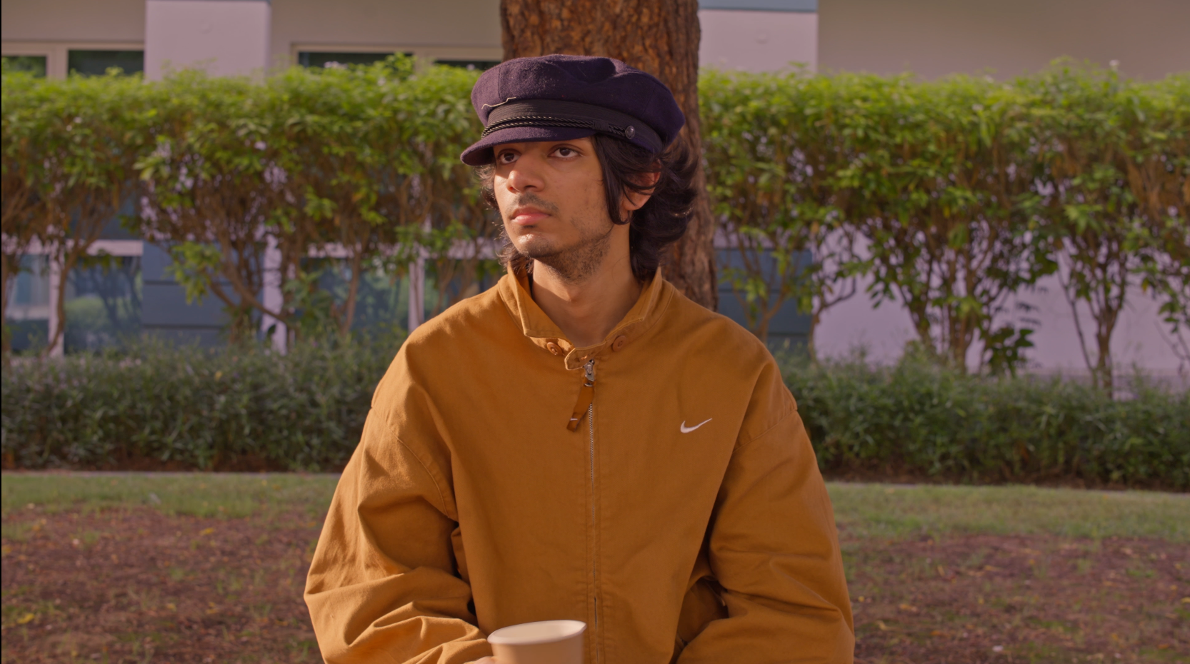A young man wearing a brown Nike jacket and a black cap sitting outdoors holding a white cup, with trees and plants in the background.