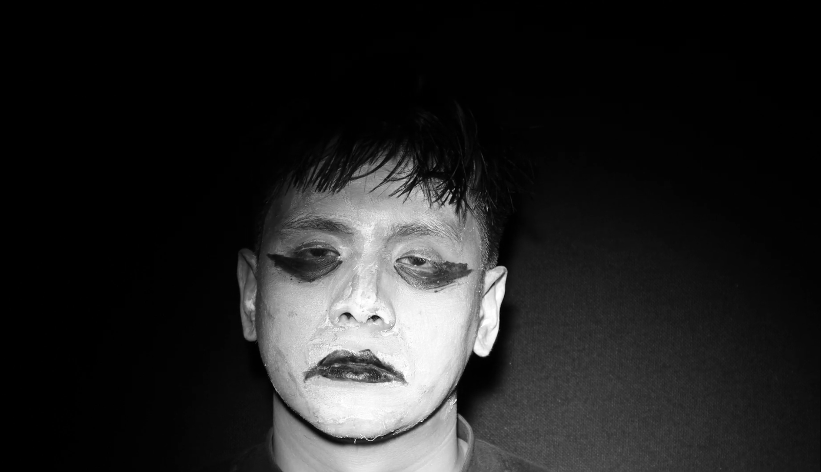 Black and white photo of a person with white face makeup, dark eye makeup, and dark lipstick, looking downward with a somber expression, against a dark background.
