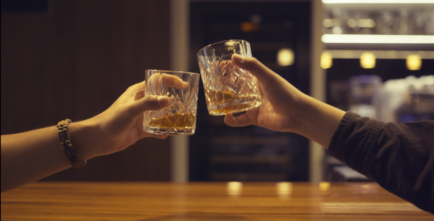 Two people clinking glasses of whiskey over a wooden bar, with a blurred background of a bar or fridge.