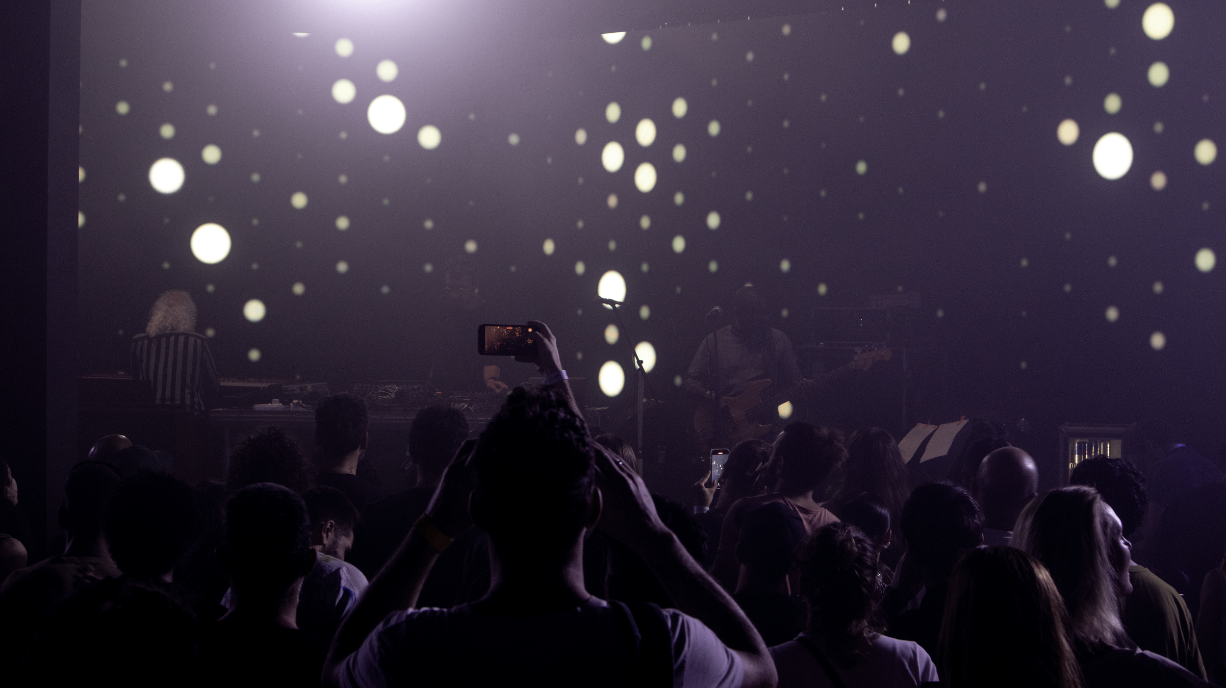 A concert scene with a crowd facing a stage where musicians perform. The background has a large screen displaying floating white dots, creating a starry or abstract pattern.
