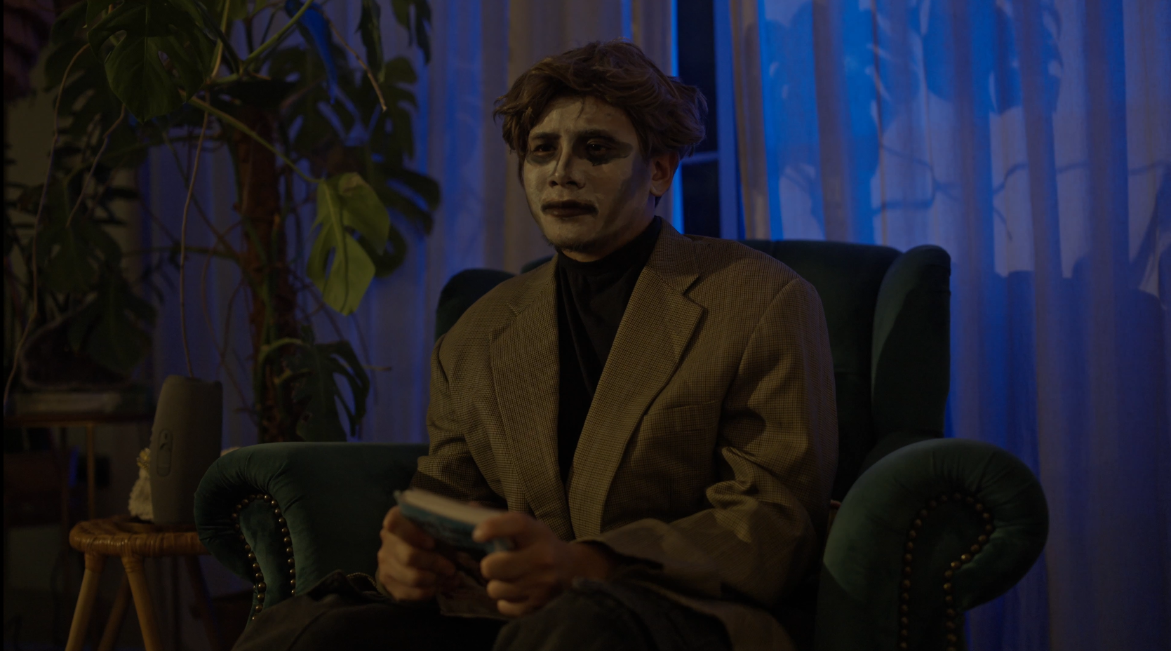 A young man with a painted face sits in a dark room with blue lighting, holding a small notebook or card. He is wearing a brown blazer over a black shirt, and appears distressed or contemplative. There is a large green plant and curtained window in t