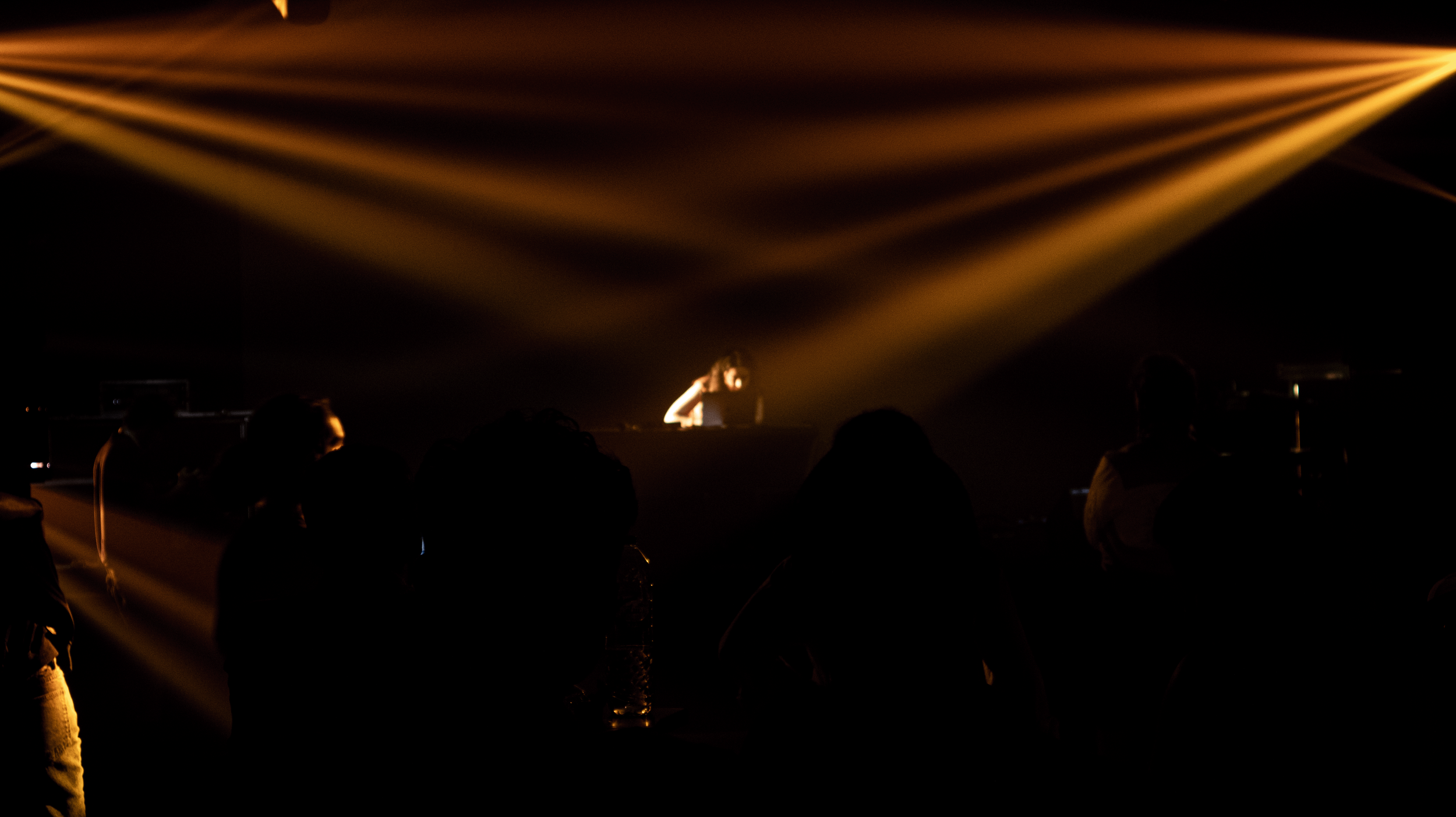 A DJ performing on stage with yellow lighting beams overhead, silhouettes of audience members watching.