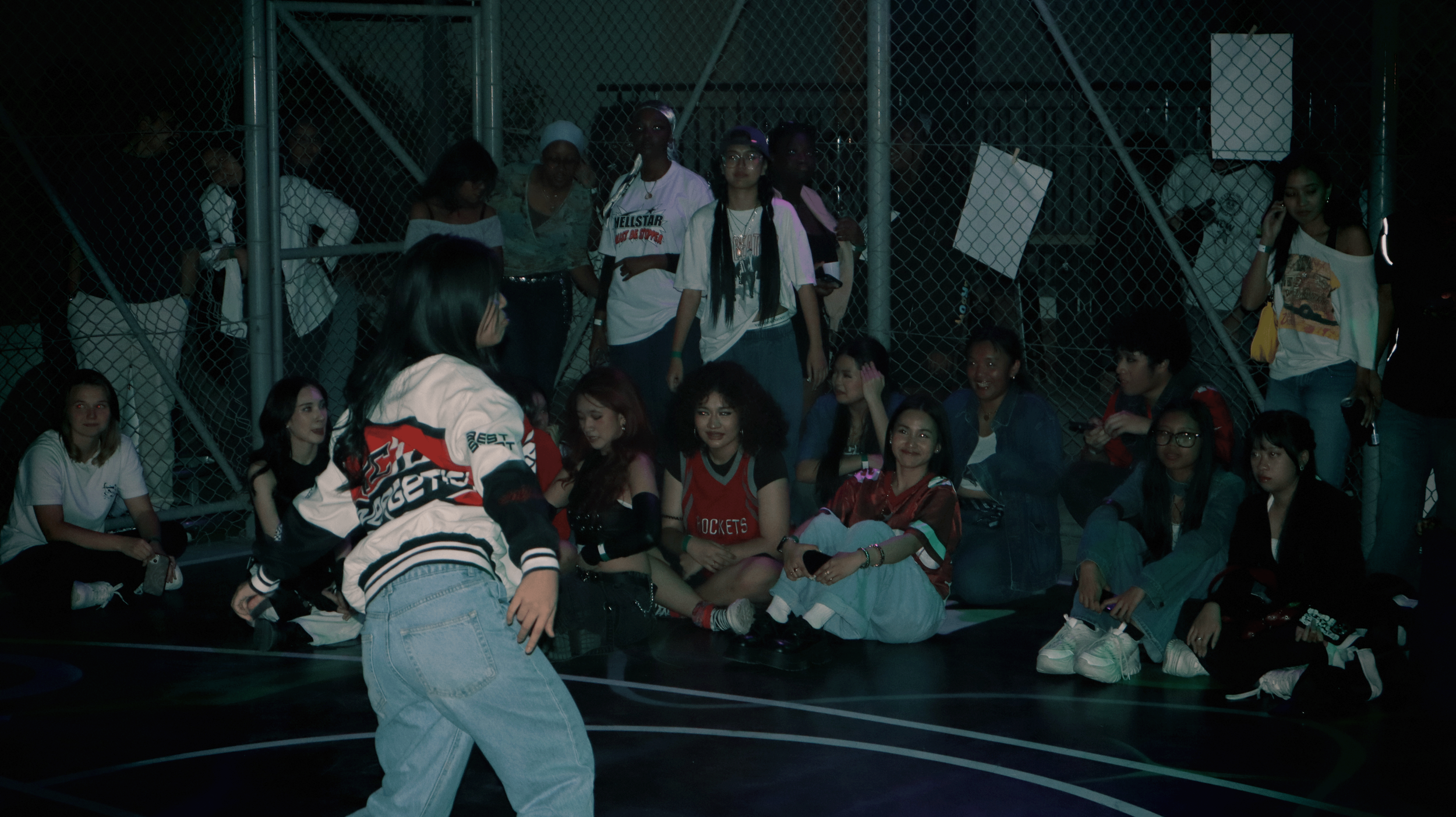 Group of young women sitting and standing behind a fence at night during an outdoor skateboarding event.