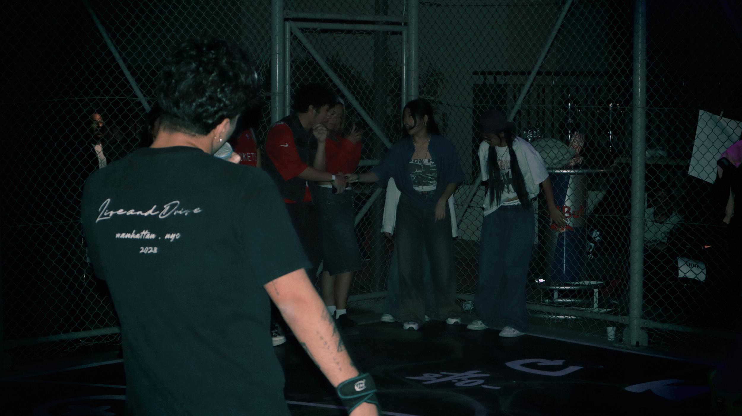 A group of young people at night on a basketball court near a chain-link fence, with one person holding a microphone, and others talking and shaking hands.