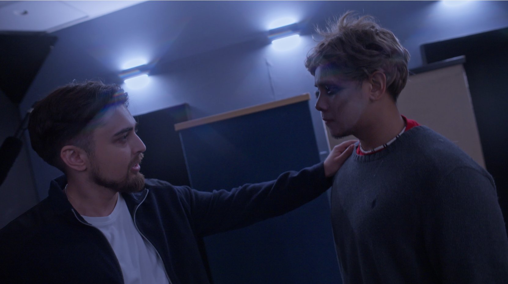 Two young men in a dimly lit room, one with a serious expression and the other with a tense look, with one placing a hand on the other's shoulder.