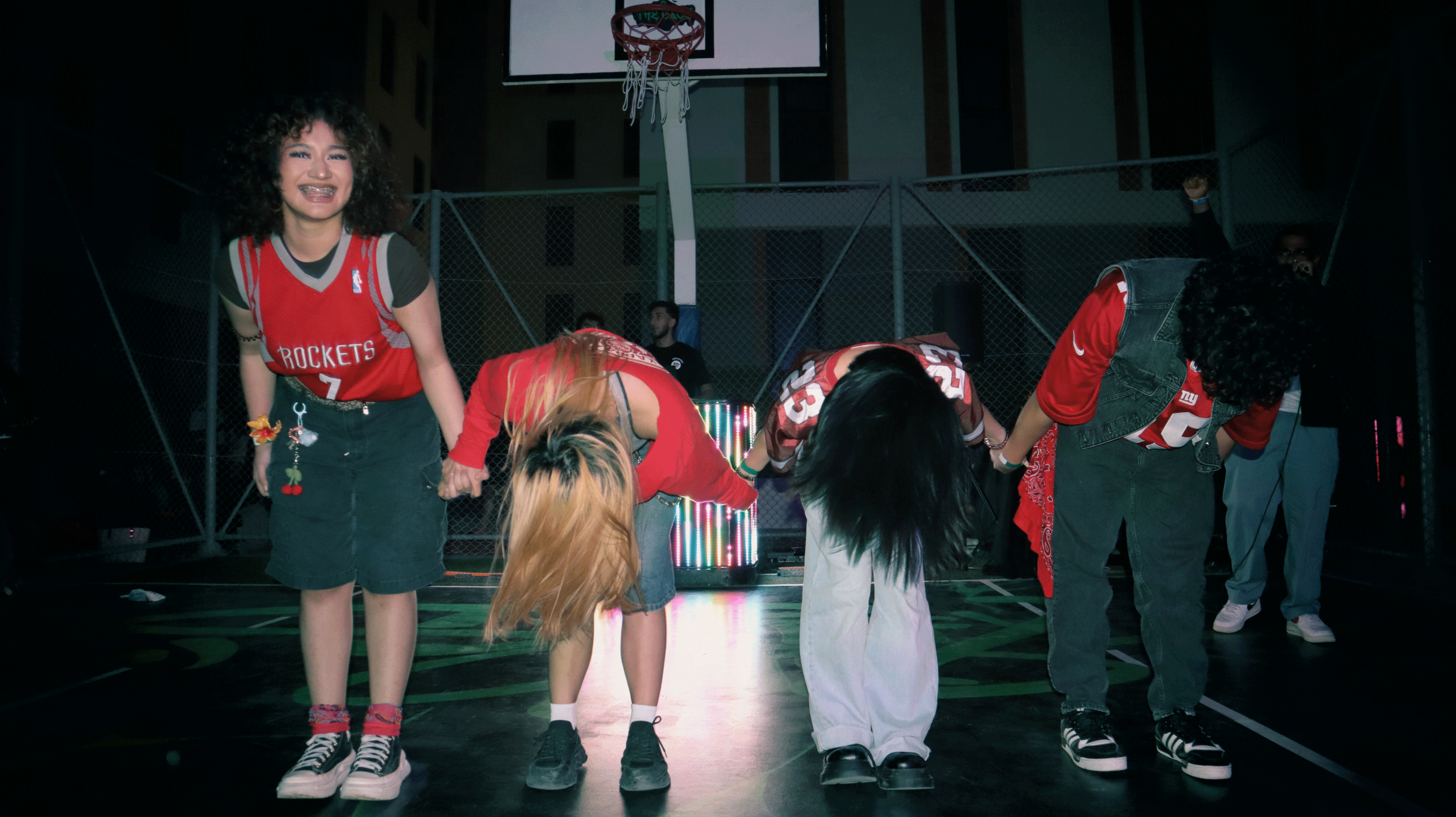 A group of young women in sporty attire, taking a bow on a basketball court at night, with a basketball hoop and a DJ setup in the background.