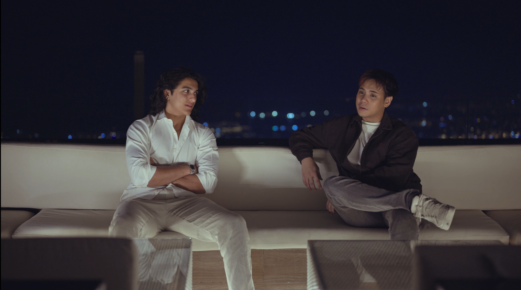 Two young men sit on a white outdoor sofa at night, having a conversation with a city skyline in the background.