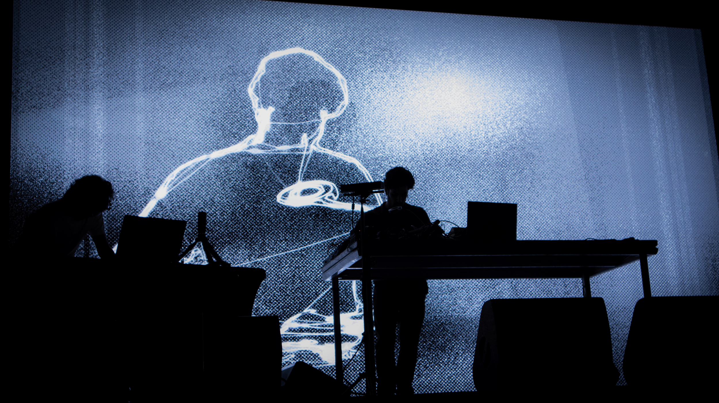 Silhouettes of two performers on stage with a large digital display in the background showing a line drawing portrait of a person wearing a cap.