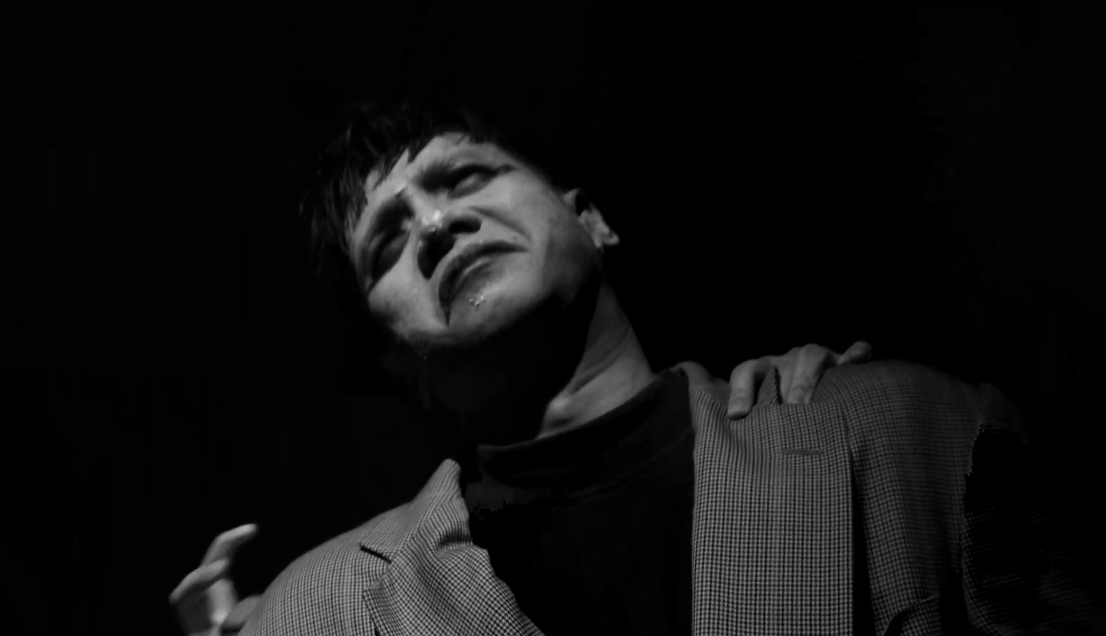 A black-and-white photo of a man with an emotional expression, one hand on his shoulder, and a hand gripping his jacket.
