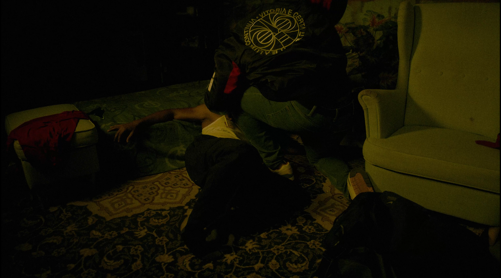 A person lying on a bed with their arm stretched out, covered with a blanket. Another person is kneeling beside the bed, reaching towards them. The room has a patterned rug, a white armchair, and various items including backpacks and clothing around.