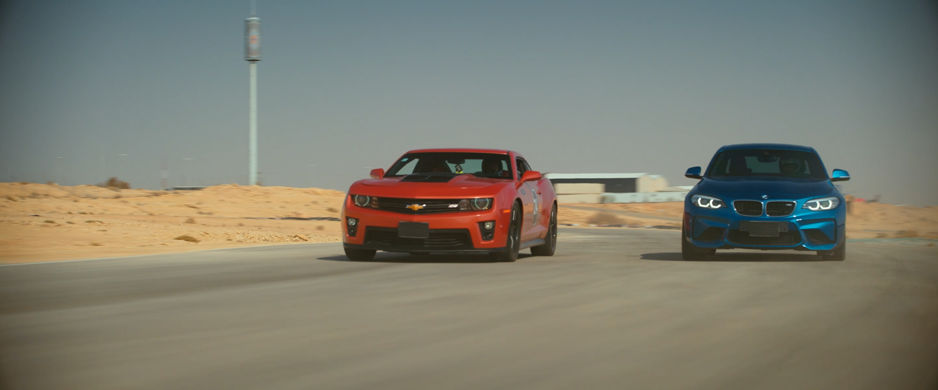 Red Chevrolet Camaro racing alongside blue BMW M2 on a desert race track.