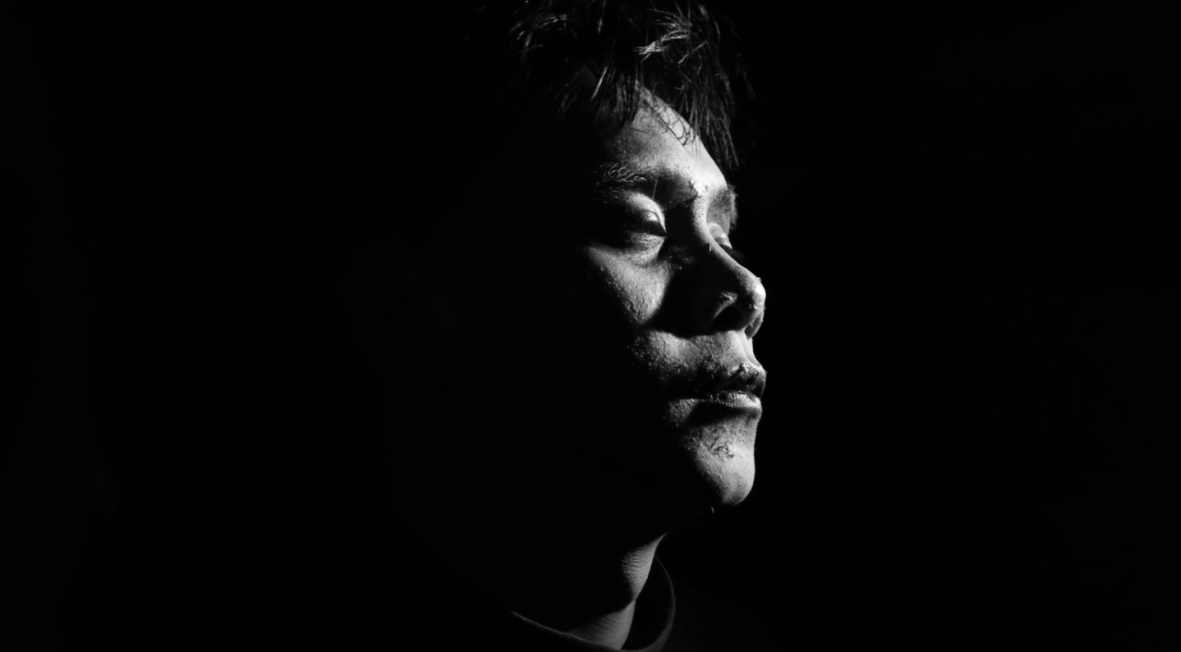 Black and white close-up of a person's face with their eyes closed, illuminated from the side, against a dark background.