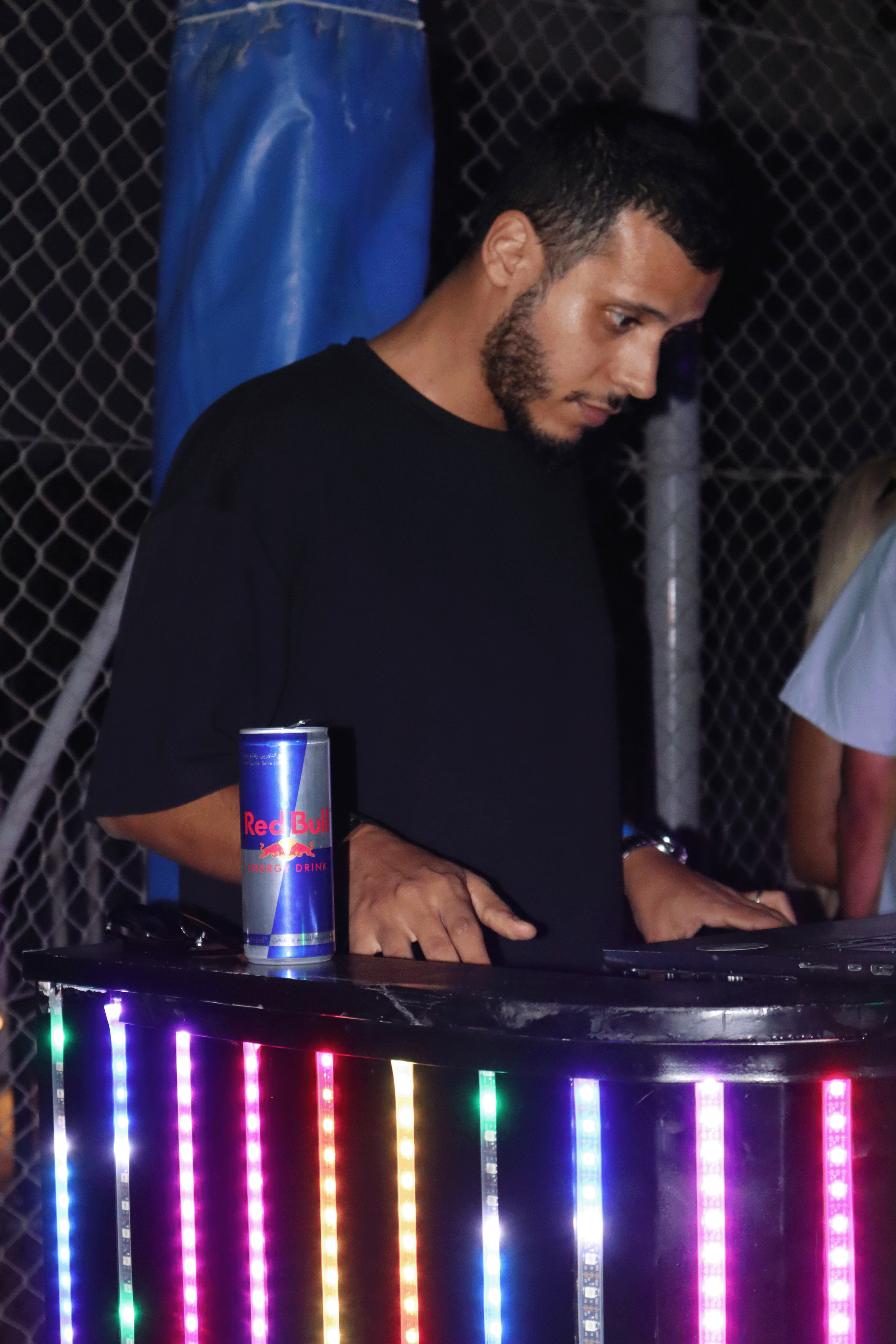 A man with short dark hair and a beard, wearing a black shirt, is standing behind a DJ booth decorated with colorful LED lights. There is a Red Bull energy drink can on the counter, and he appears to be focused on playing music. The background featur