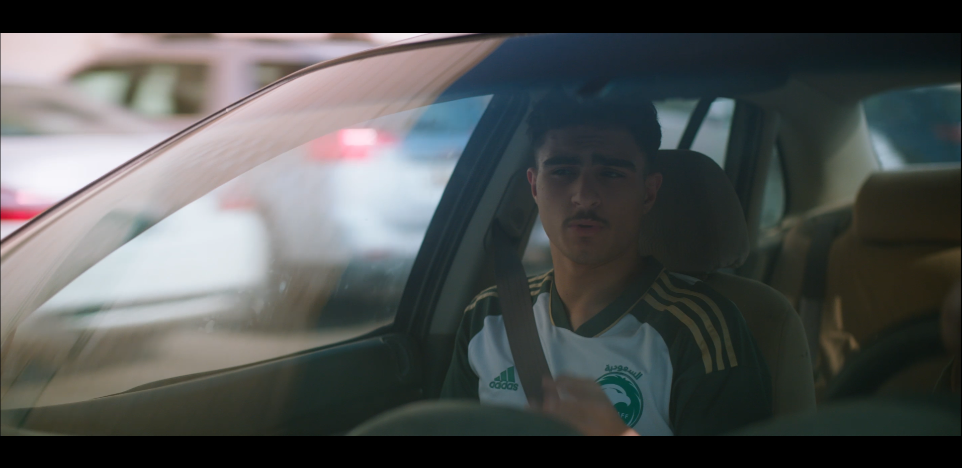 A young man sitting in the driver's seat of a car, wearing a Saudi Arabia national football team jersey, with a focused expression, looking through the car window.