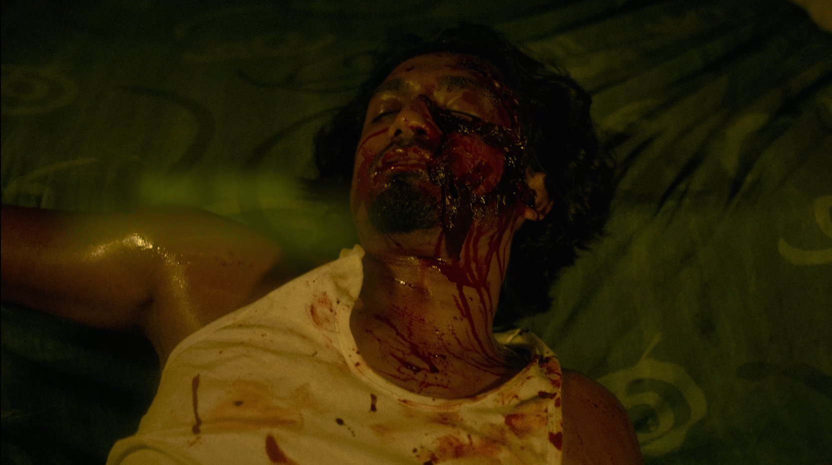 A person lying on a bed with blood on their face, neck, and shirt, appearing to be injured or dead.