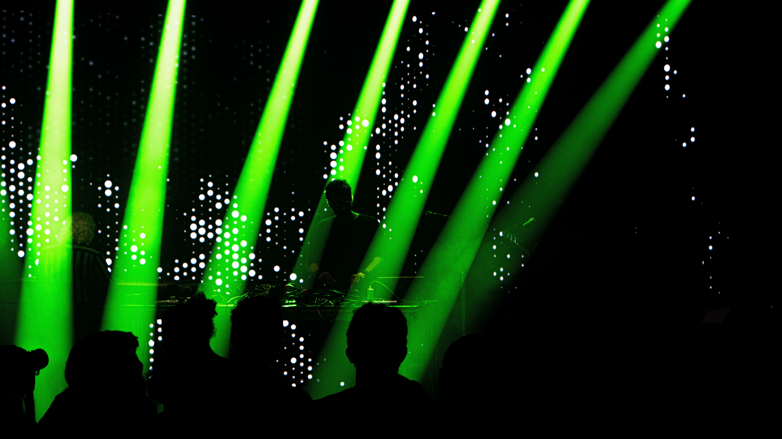 Silhouettes of people watching a concert with green stage lights and a performer on stage with digital background visuals.